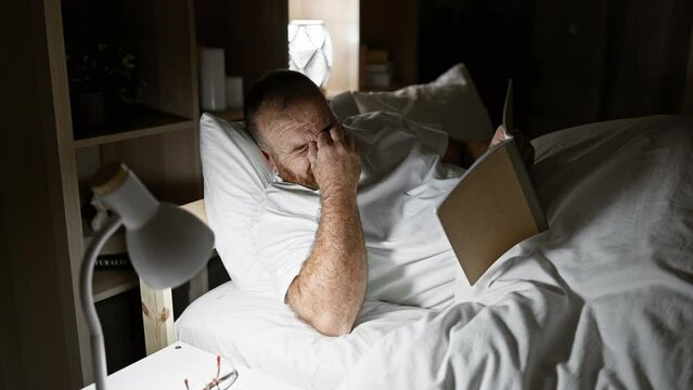 Attractive, bearded caucasian man lying comfortably on bed in lamp-lit bedroom, indulging in morning leisure by reading book, wearing glasses - a portrait of relaxed indoor lifestyle.