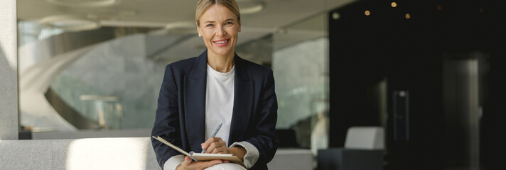 Smiling female manager making notes in note pad sitting on modern office background