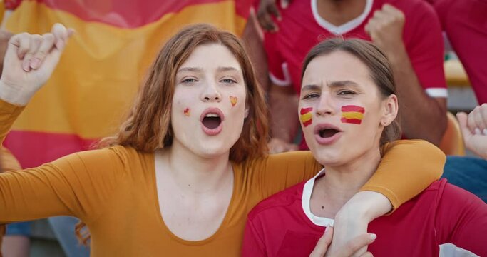 Couple of pretty Caucasian women with Spanish national flags painted on their cheeks. Girls hugging each other and singing something at stadium tribunes to support their favourite team.