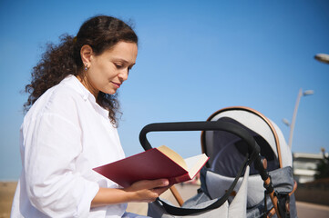 Happy young Latina woman, mother pushing baby stroller, reading book, sitting relaxed on the...