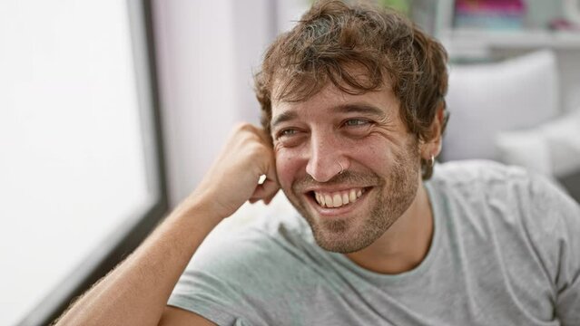 Handsome young man, chilling at home, confidently smiling and looking away. portrait of hispanic guy with beard, sitting on sofa, laughing naturally in cozy living room.