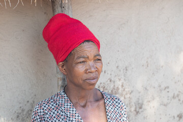 old African woman with red scarf, natural light against the wall of the mud house in the yard of...