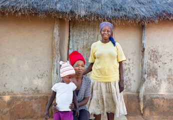 village african family three generations , standing in the yard daytime, in front of the thatched...