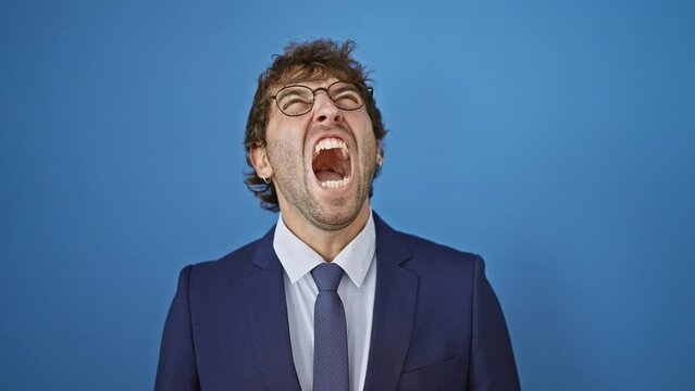 Frustrated young man in business suit, seething with anger and aggression, screams out his fury. a stark portrait of rage, isolated on a blue background.