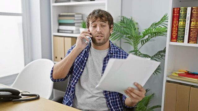 Captivating portrait of a hard-working young man, reading a business document while engaging in a phone conversation at his office desk