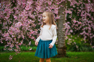 Adorable preschooler girl enjoying nice spring day in park during cherry blossom season