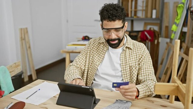 Handsome bearded man shopping online with credit card on tablet at carpentry workshop