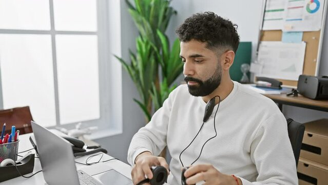 A young bearded man wearing a headset looks troubled while working at his laptop in a modern office setting.