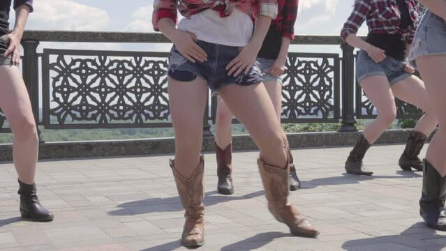 Several young girls dancing country line dance on the asphalt, USA style. Close up view to female hips and feet with brown cowboy country boots and jeans shorts. A little slow motion video