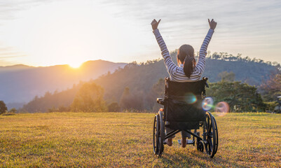 Happy person sitting on a wheelchair enjoying with raised hands on the mountain at the sunset time.