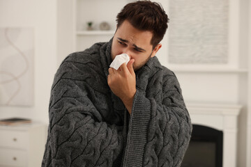 Sick man wrapped in blanket with tissue blowing nose at home. Cold symptoms