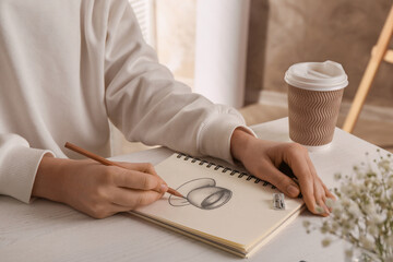Woman drawing cup of drink with graphite pencil in sketchbook at table, closeup