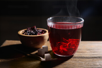 Delicious hibiscus tea in glass, sugar cubes and dry roselle petals on wooden table, closeup