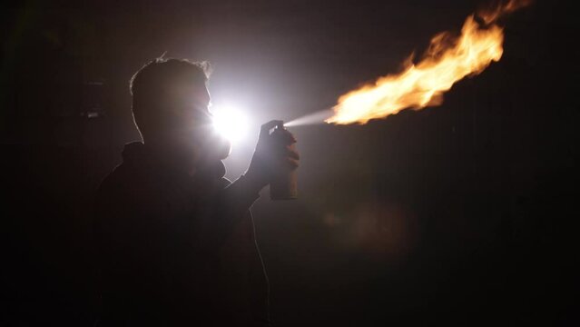 Fire demonstration, Protective mask, Flame performance. Amidst darkness and haze, silhouette skillfully uses aerosol can to produce vivid flame, protected by safety mask.