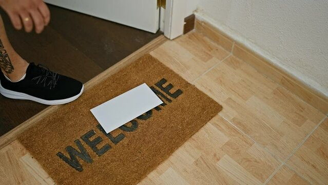 A tattooed man in a beige shirt reacts with apprehension to an envelope on a 'welcome' doormat in a home hallway.