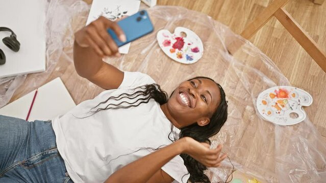 A cheerful young woman with curly hair takes a selfie while lying on a floor with art supplies around her indoors.