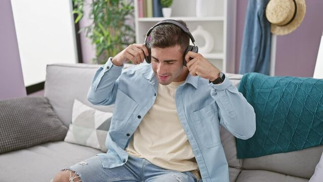Handsome young hispanic man, relaxed on sofa, joyously dancing to the rhythm of music he's listening to via hi-tech gadget at home