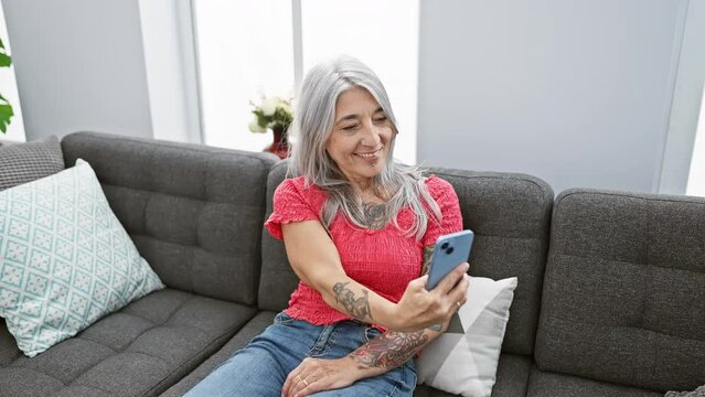 Joyful mature woman enjoying her time at home, happily taking a selfie sitting on her cosy sofa, with a beaming expression and her grey hair as her phone's background