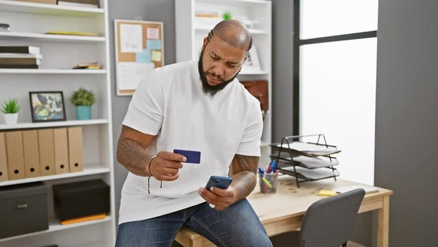 African american man with beard checks credit card and smartphone in modern office, expressing focused curiosity.