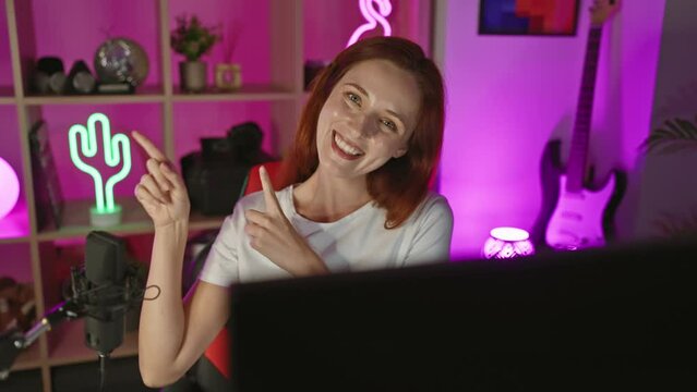 Cheerful young redhead caucasian woman streamer looking at camera, smiling and pointing to side in gaming room at home office night