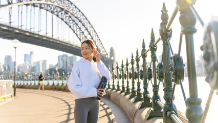 Happy Asian woman listening to music from mobile phone and earphones with mobile app during jogging exercise at city street in the morning. Healthy girl do sport training running workout in the city. - obrazy, fototapety, plakaty