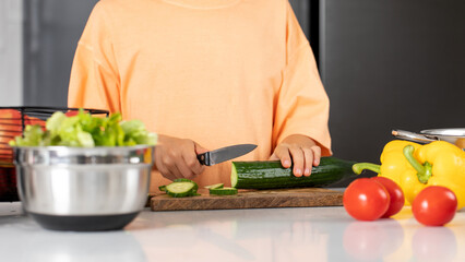 A boy is cutting a cucumber at the table. The child is preparing a vegetable salad. Healthy food