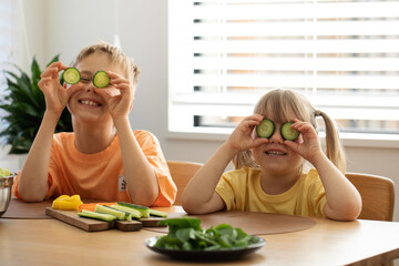 Funny brother and sister are sitting at the table and funny holding cucumbers to their eyes.