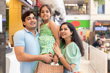 Cheerful Indian family of three together with paper bags, shopper bags in mall, father and mother holding their daughter. Shopping concept