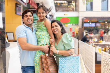 Cheerful Indian family of three together with paper bags, shopper bags in mall, father and mother holding their daughter. Shopping concept