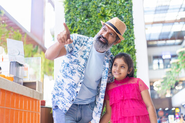 Senior looking Indian father wearing hat point aside with daughter and enjoying in shopping mall.