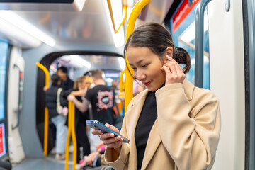 Asian woman using mobile phone and listening to the music on earphones during travel on tram in the city. Attractive girl enjoy urban lifestyle in the city with using wireless technology on device. - obrazy, fototapety, plakaty
