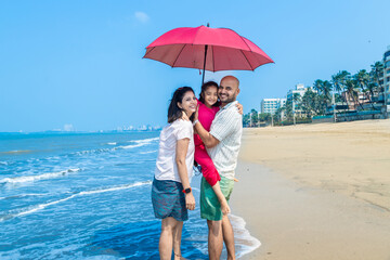 Happy indian family of three standing under sun umbrella enjoying summer vacation on tropical beach. Father holding her daughter on beach.