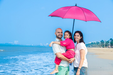 Happy indian family of three standing under sun umbrella enjoying summer vacation on tropical...