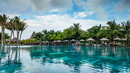 Tropical resort swimming pool with palm trees, sun loungers, and thatched huts under a cloudy sky,...