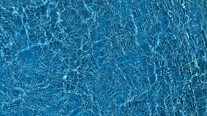 A close-up texture of sunlit, clear blue swimming pool water rippling gently, ideal for summer or...