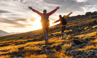 Couple hikers are walking together in sunset mountains. Two young tourists with backpacks are standing with open arms and enjoys sunset - obrazy, fototapety, plakaty