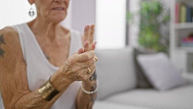 Senior grey-haired woman suffering intense hand pain, sitting in living room sofa at home, elderly female with short hair looking hurt, osteoarthritis problem indoors.