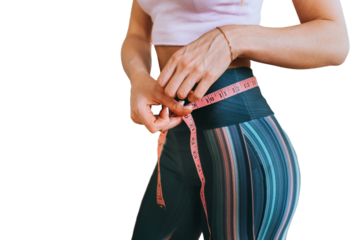Cropped image of fit caucasian young woman measuring waist by measuring tape stands against transparent background against blurry dumbbells. Weight loss concept.