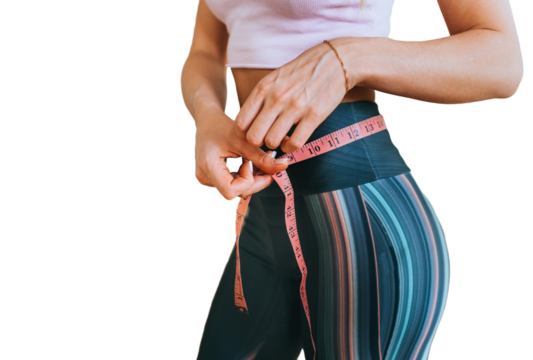 Cropped image of fit caucasian young woman measuring waist by measuring tape stands against transparent background against blurry dumbbells. Weight loss concept.