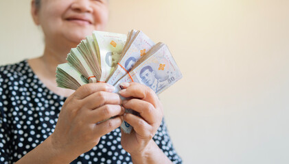 Happy older Asian woman holding a lot of Thai money that she has won, feeling excited.