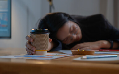 Overworked and tired businesswoman sleeping while hand over a cup of coffe in a desk at work in her...