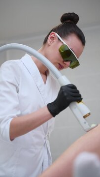 Skilled aesthetician performs laser hair removal in clinic, patient undergoing epilation treatment. Focused pro wields advanced device, ensures comfort during depilation session. Safe body care.