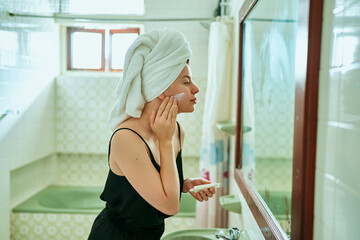 Woman in a vintage bathroom applies sunscreen to her face for skin protection. Towel-wrapped hair,...