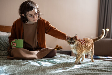 Loving teen girl sitting on bed with cup of tea, stroking, caressing breed cat Devon Rex. Pets...