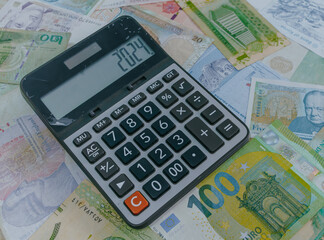2024 numbers on a calculator and background of banknotes from different countries. New year finance, money inflation and budget concept.
