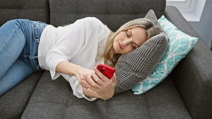 A young blonde woman in casual attire relaxes on a sofa, using a smartphone before dozing off in a cozy living room. - Powered by Adobe