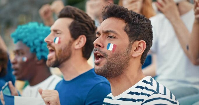 Couple of multi-ethnic people with French flag painted on their faces. Shouting words of support while spectating game from stadium tribunes. Shaking French flag to show their support.