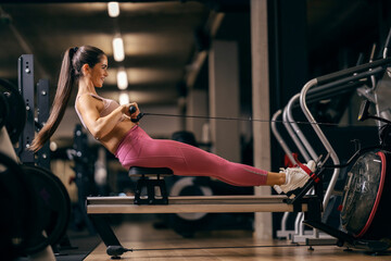 A strong sportswoman is doing exercises on rowing machine.