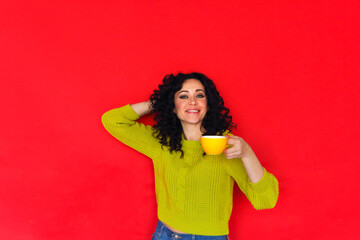 Young happy curly woman with cup of tea smiling at camera while posing on red studio background