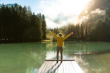 Back of young man with hands raised standing on a wooden pier and enjoying the scenery. Adventure travel in Slovenia, mountain lake in Jezersko. Vibrant autumn colors on a sunny day in the Alps.